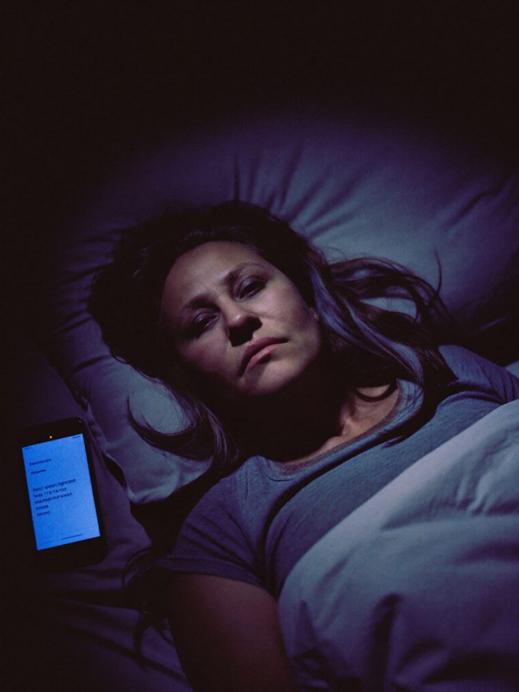 A woman lying awake in bed at night, lit by a phone screen